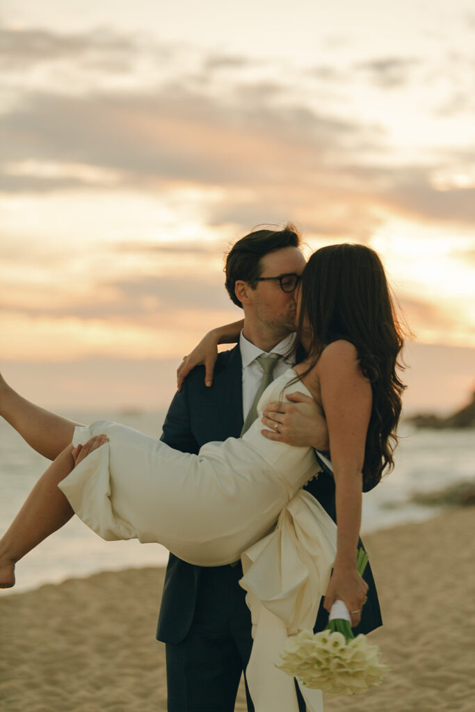 groom carries bride on beach at Mexico sunset destination wedding
