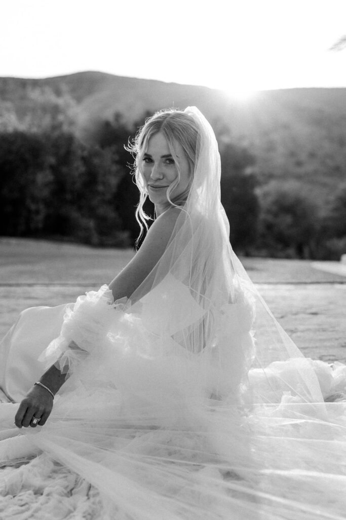 bride smiling with cape and veil sitting on the ground