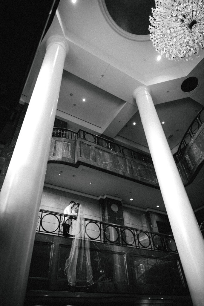 bride and groom leaned over the railing with view of entire hotel room and chandelier
