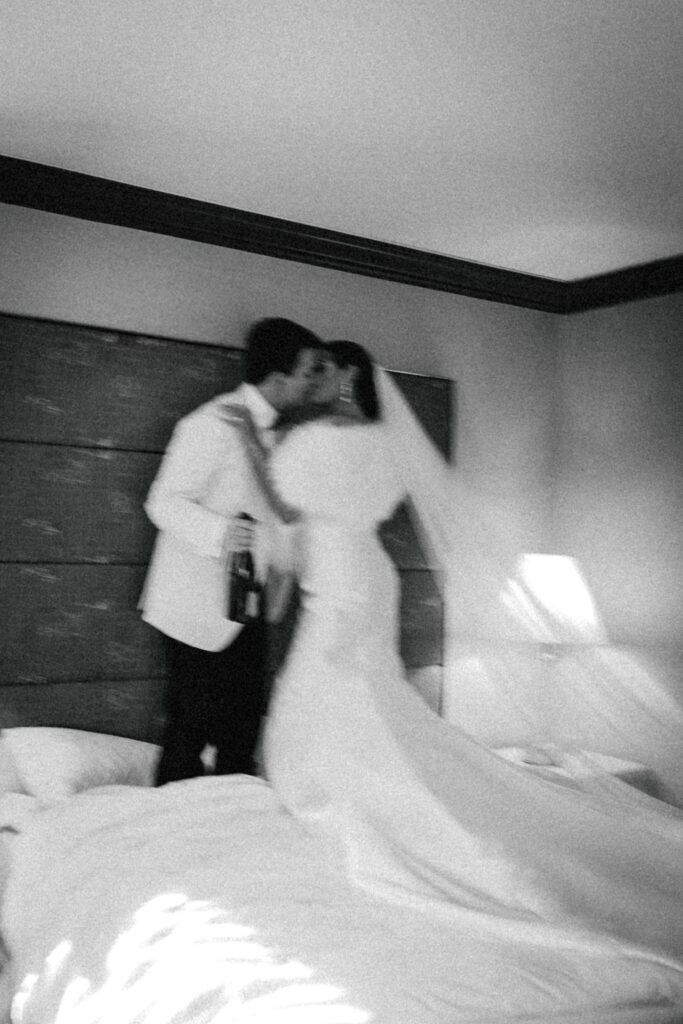 bride and groom kissing and jumping on hotel bed at wedding venue