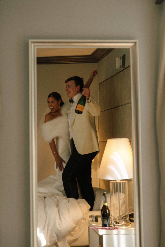 bride and groom in mirror jumping on hotel room bed with champagne