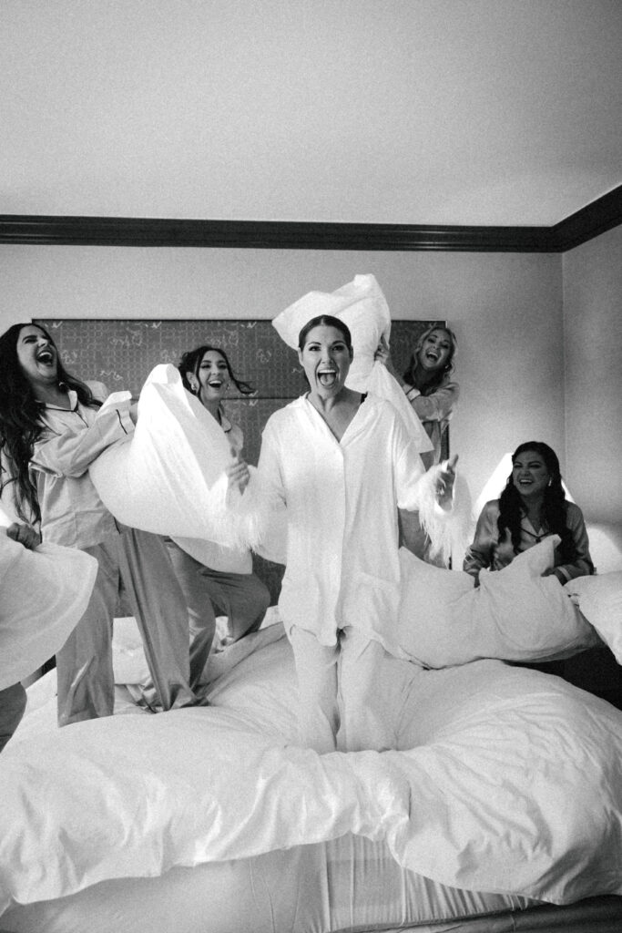 bride and bridesmaids pillow fight on wedding bed in hotel room