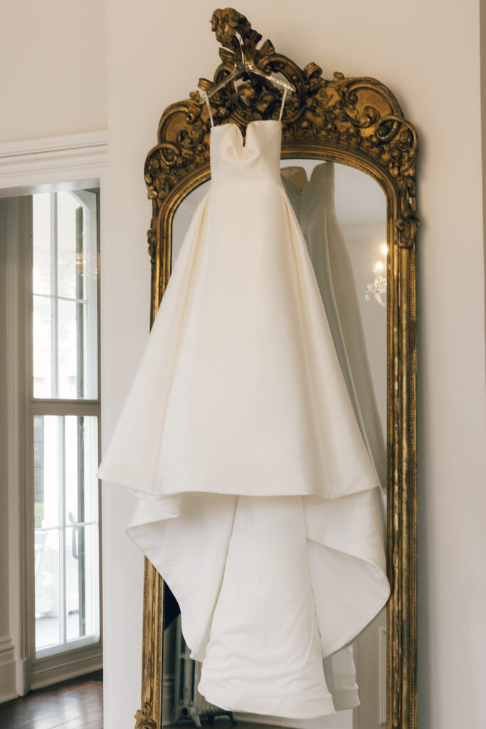 white wedding dress hangs over grand gold mirror 